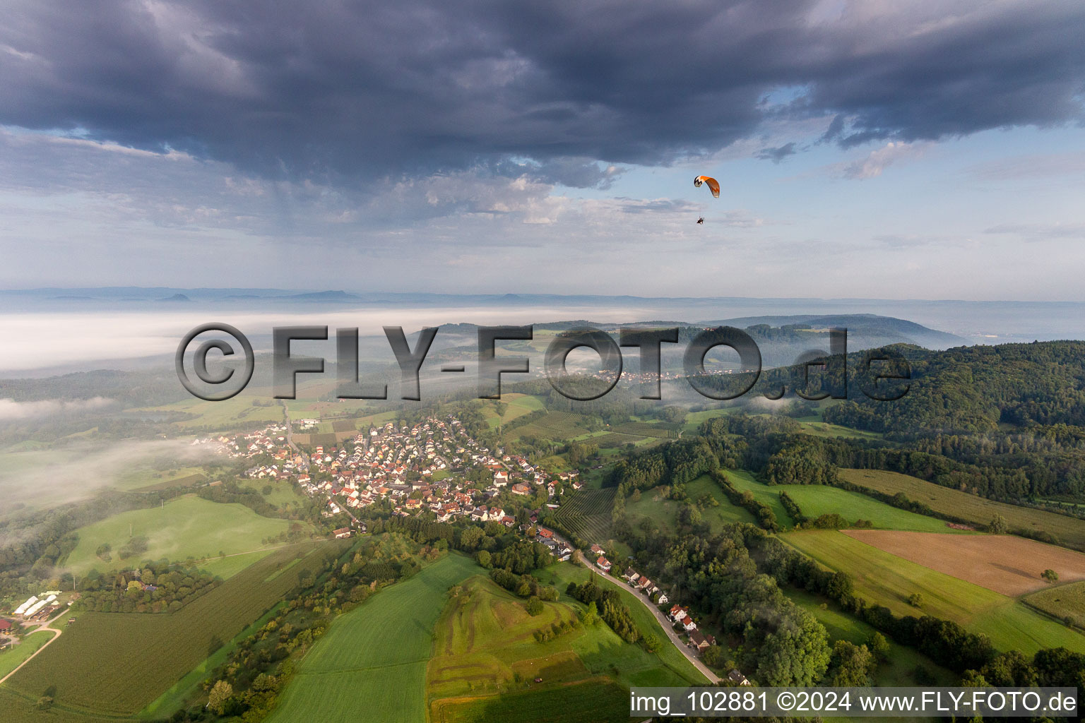 Paraglider before the Lake of Constance hidden by mroning fog over Village - view on the edge of agricultural fields and farmland in the district Guettingen in Radolfzell am Bodensee in the state Baden-Wurttemberg, Germany